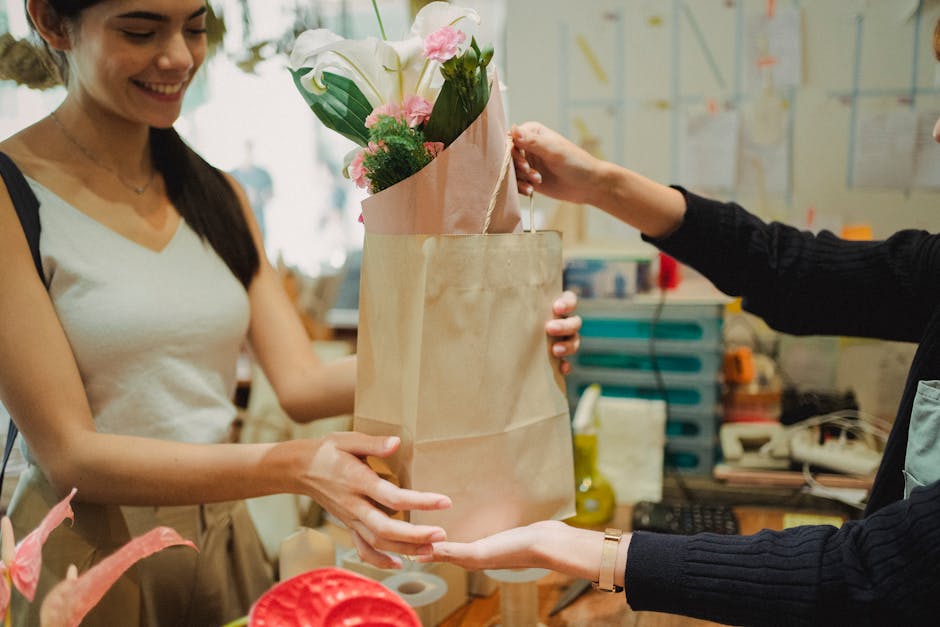 A woman in a white sleeveless top is smiling as she receives a bouquet of fresh flowers, including pink roses and white lilies, wrapped in pink and brown paper. She holds a brown paper gift bag, and a