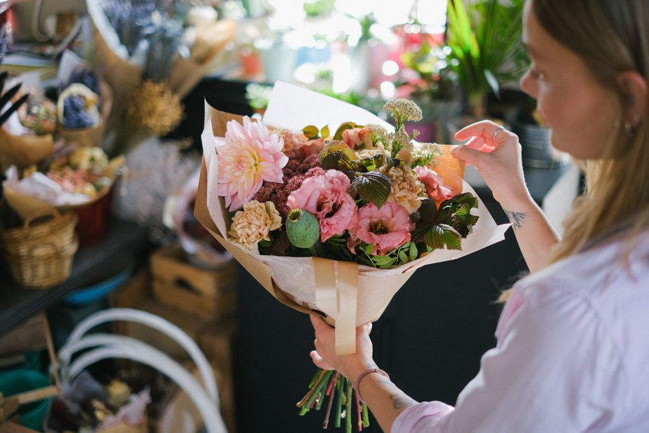 A woman with light brown hair, wearing a light pink shirt, holds a large bouquet of fresh flowers in a florist shop. The bouquet includes pink dahlias, light pink roses, and green foliage, arranged in