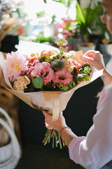 A person holding a large bouquet of fresh, vibrant pink and cream flowers, including peonies, dahlias, and carnations, wrapped in cream and white tissue paper with a beige ribbon. The arrangement feat