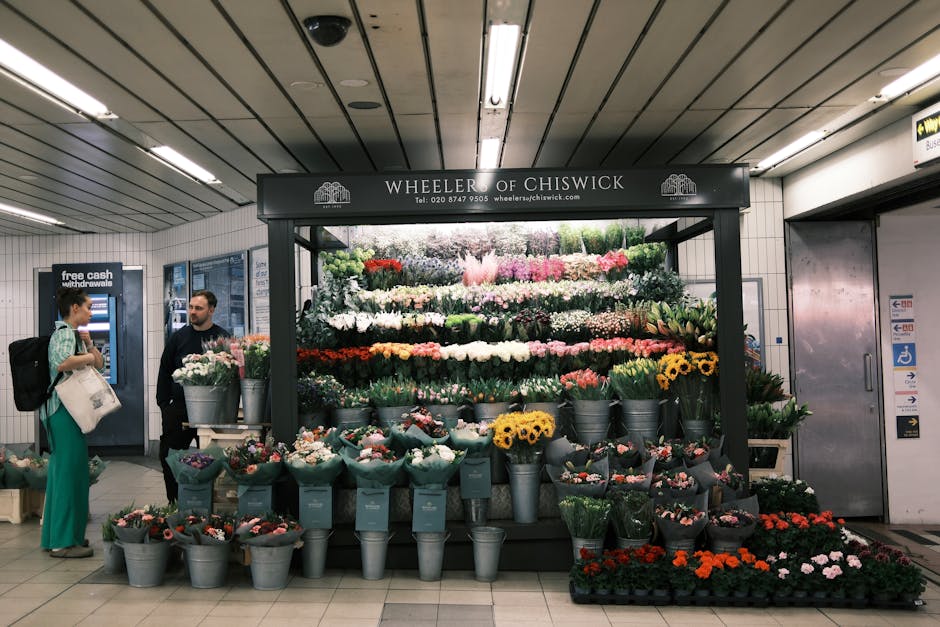 A floral display stand at Wheeler of Chiswick inside a train station, featuring a variety of fresh flowers arranged in buckets and bouquets. The arrangement includes prominent pink, white, yellow, red